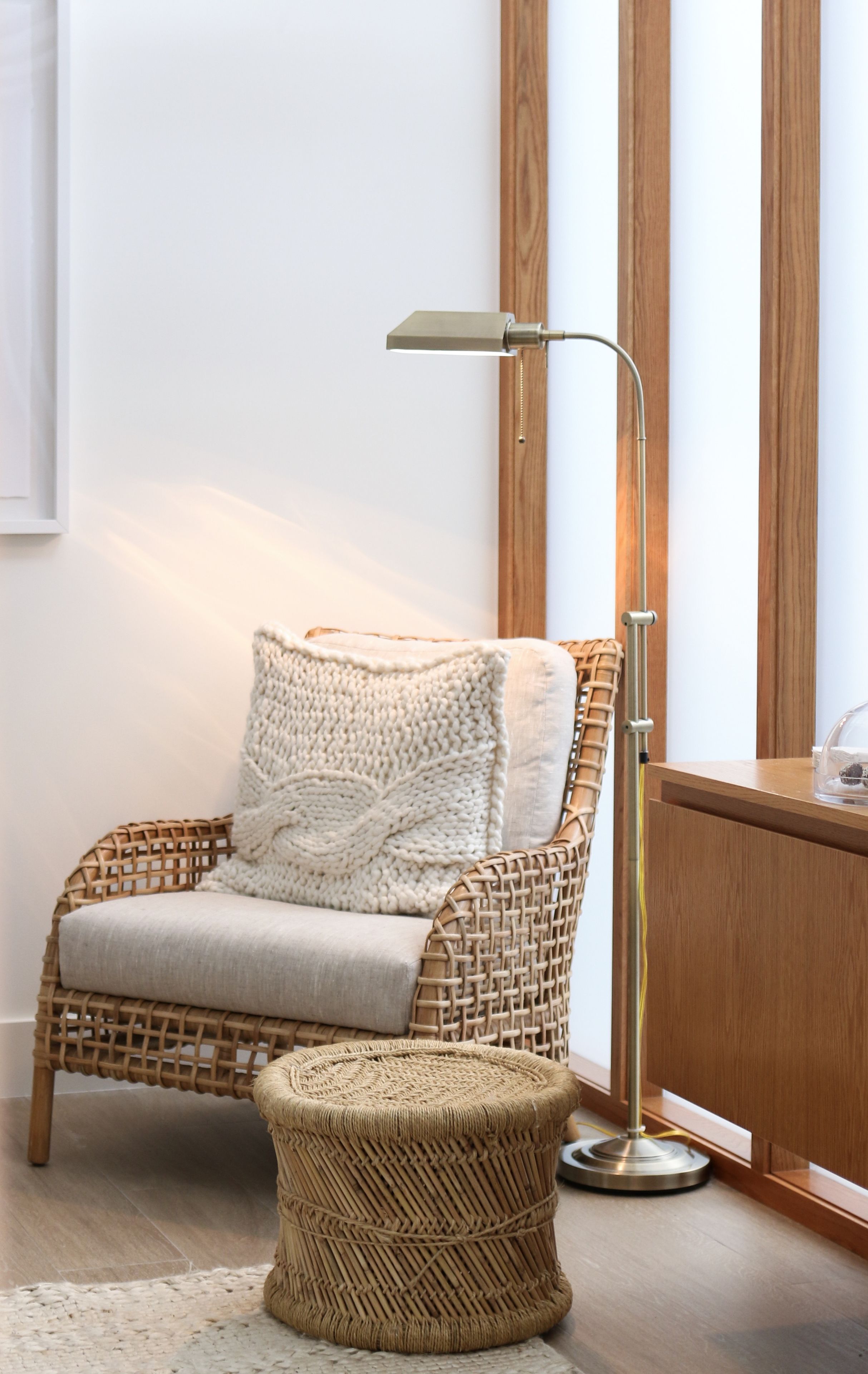 Een rieten stoel met kussen, een klein geweven krukje en een vloerlamp in een kamer met houten lambrisering en een witte muur.