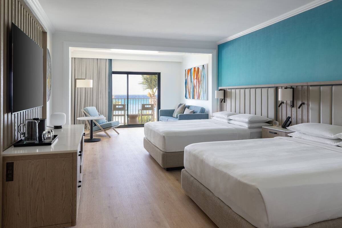 A hotel room with two beds, a desk with a coffee maker, a TV, and a balcony with a view.