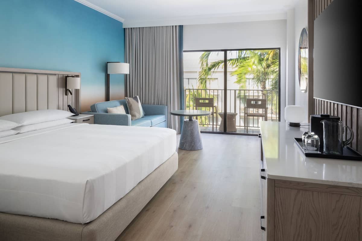 A hotel room with a large bed, a seating area with a table and chairs, a balcony with a view of palm trees, and a kitchen area with a coffee maker.