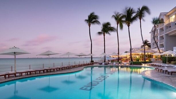 A poolside area with lounge chairs and umbrellas, a clear view of the ocean, and a building with balconies. - Grand Cayman Marriott Resort (Seven Mile Beach)