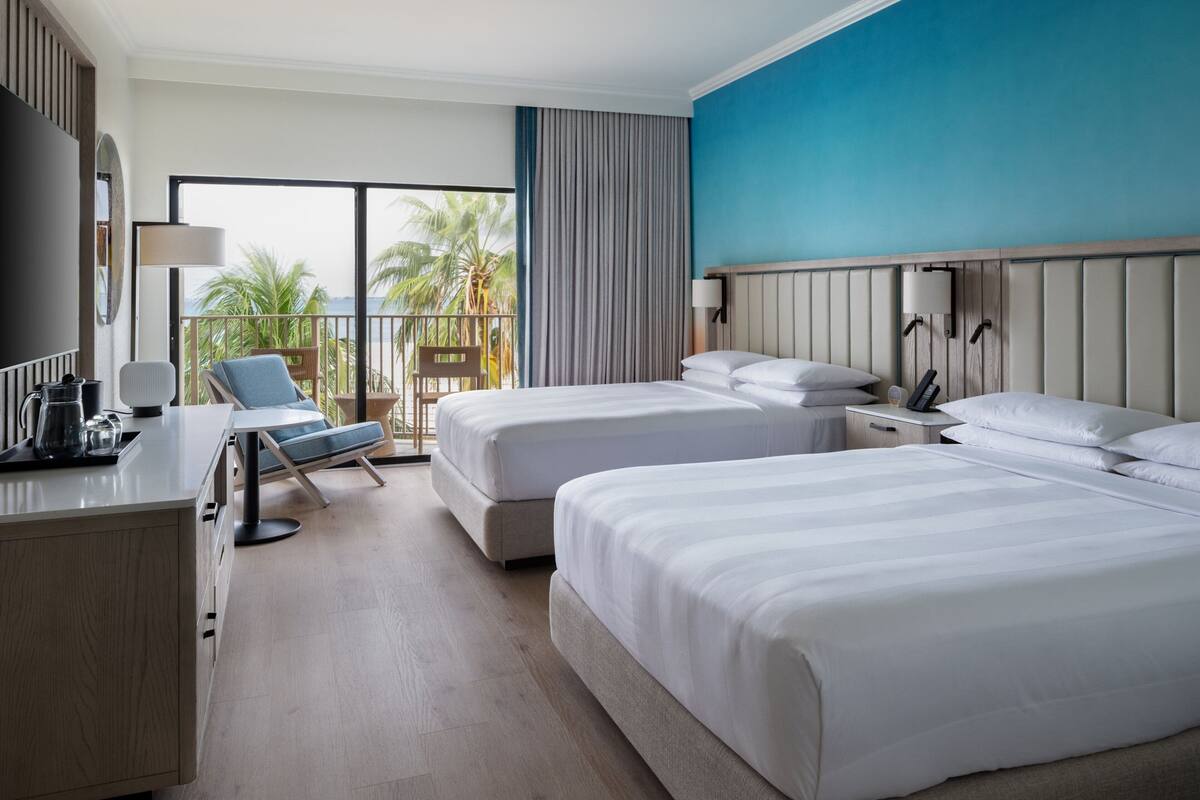 A hotel room with two beds, a desk with a lamp, a chair, and a view of palm trees through large windows.