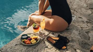 Una mujer sentada junto a una piscina con una bebida y aperitivos, rodeada de sillas de descanso y la piscina.