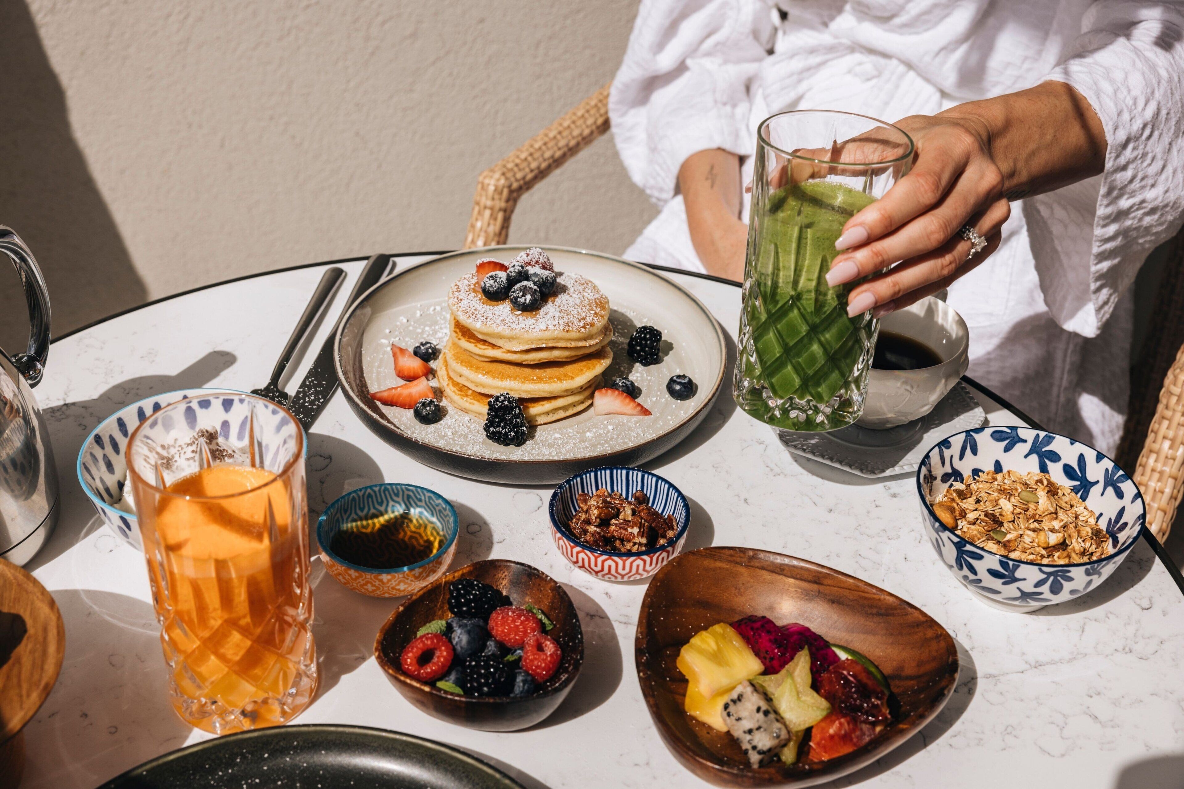 Un desayuno con tortitas, frutas y bebidas sobre una mesa.