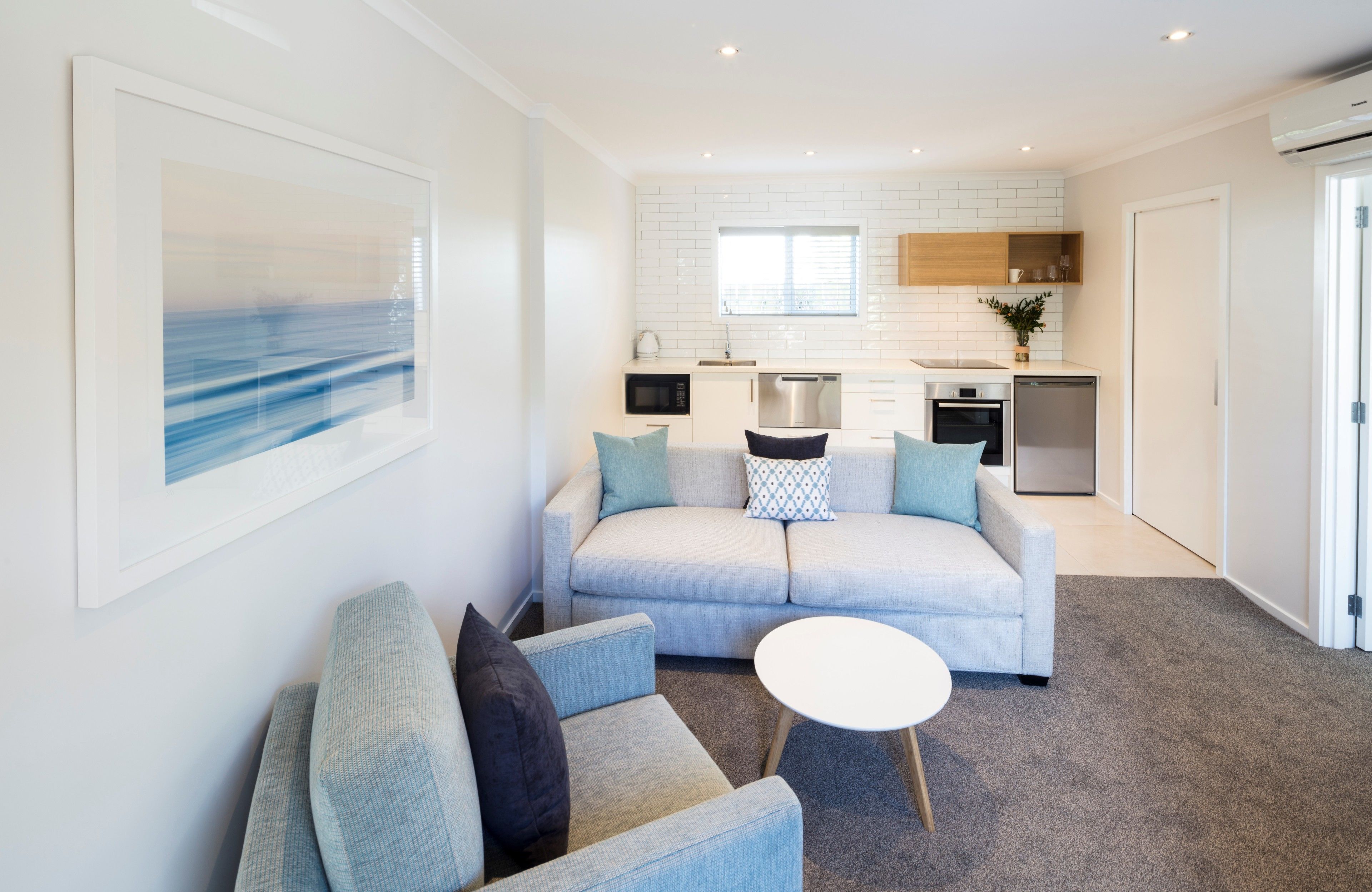 A modern living room with a sofa, armchairs, and a coffee table. A kitchen with white appliances and a window is visible in the background.