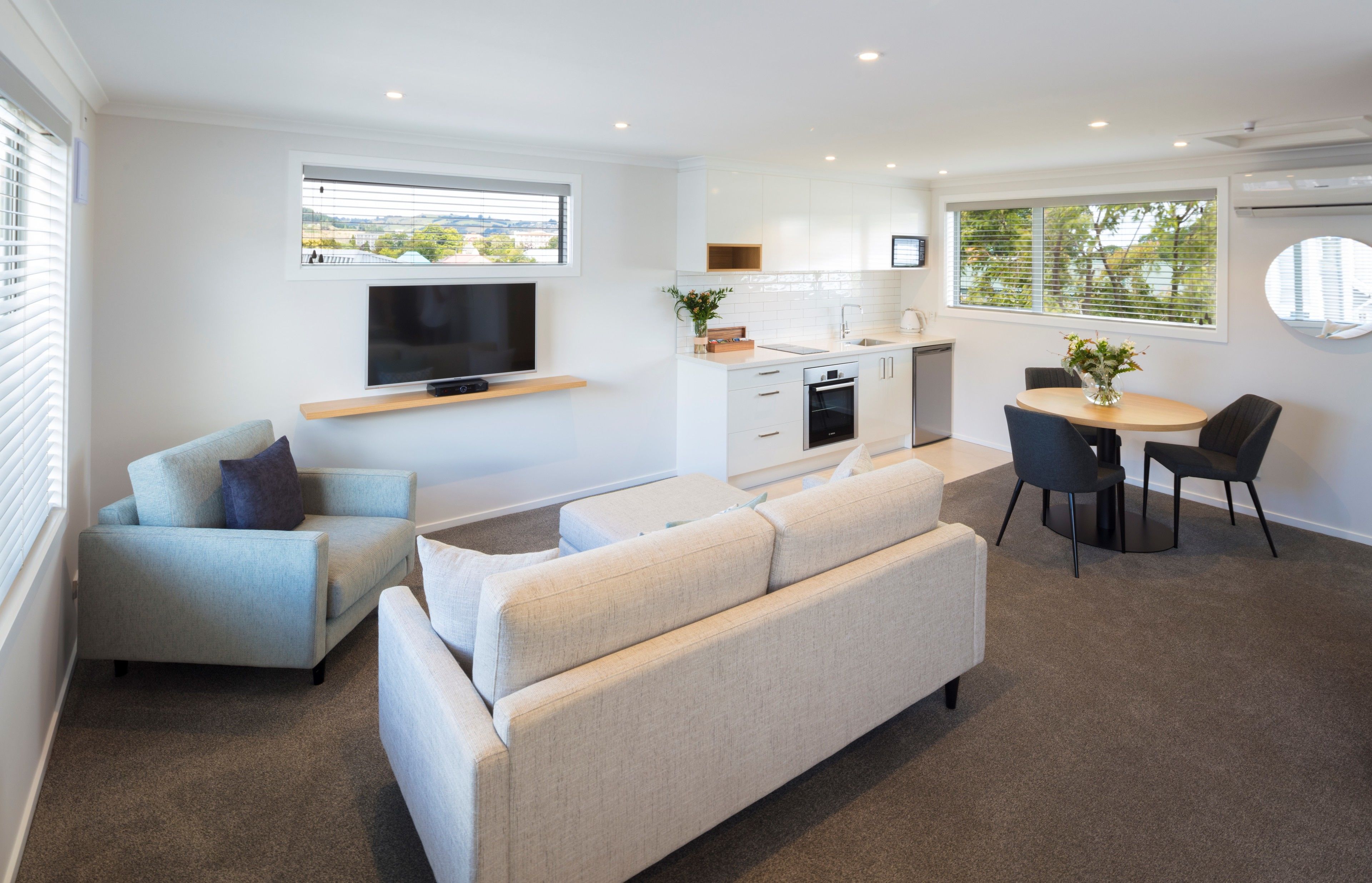A modern living room with a sofa, a TV mounted on the wall, a dining area with a table and chairs, and a kitchen with cabinets and a window.