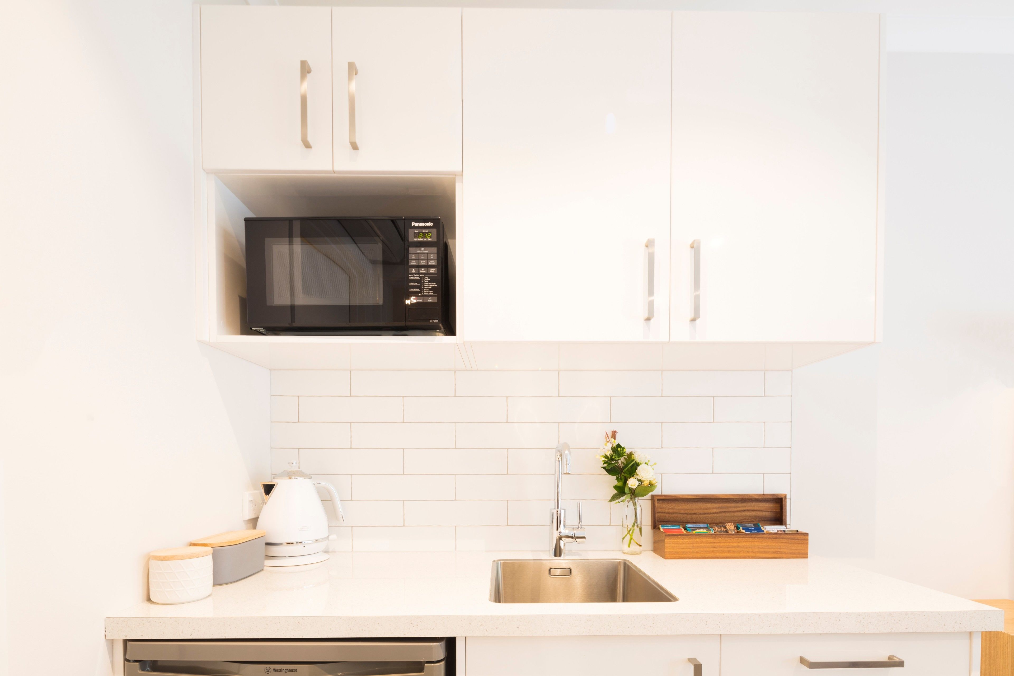 A modern kitchen with white cabinetry, a built-in microwave, a stainless steel sink, and a coffee maker.