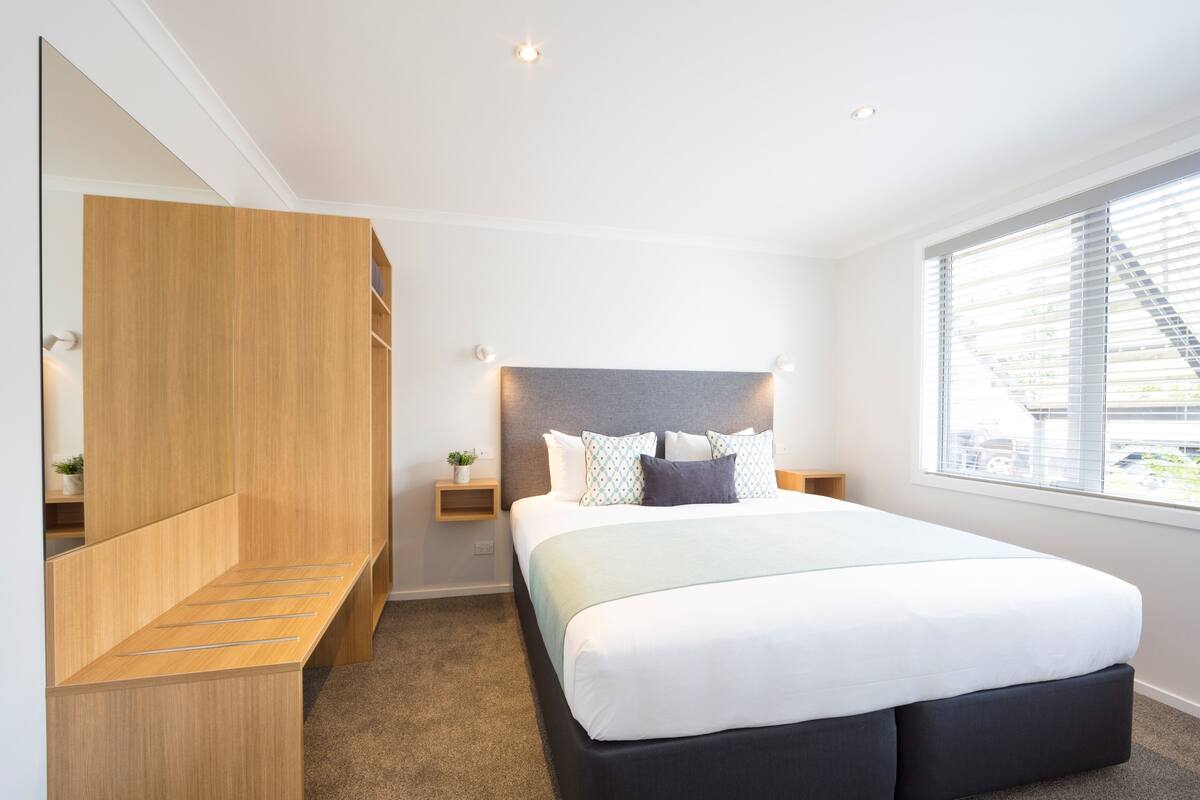 Un dormitorio moderno con una cama grande, armario de madera empotrado, mesitas de noche y una ventana con persianas.