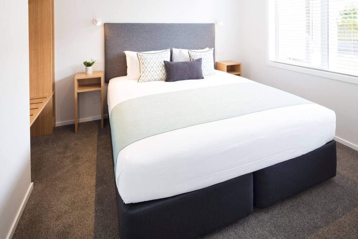 Un dormitorio moderno con una cama grande, un cabecero gris, una mesita de noche con una lámpara y una ventana con persianas.