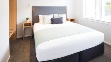 A modern bedroom with a large bed, a grey headboard, a bedside table with a lamp, and a window with blinds.