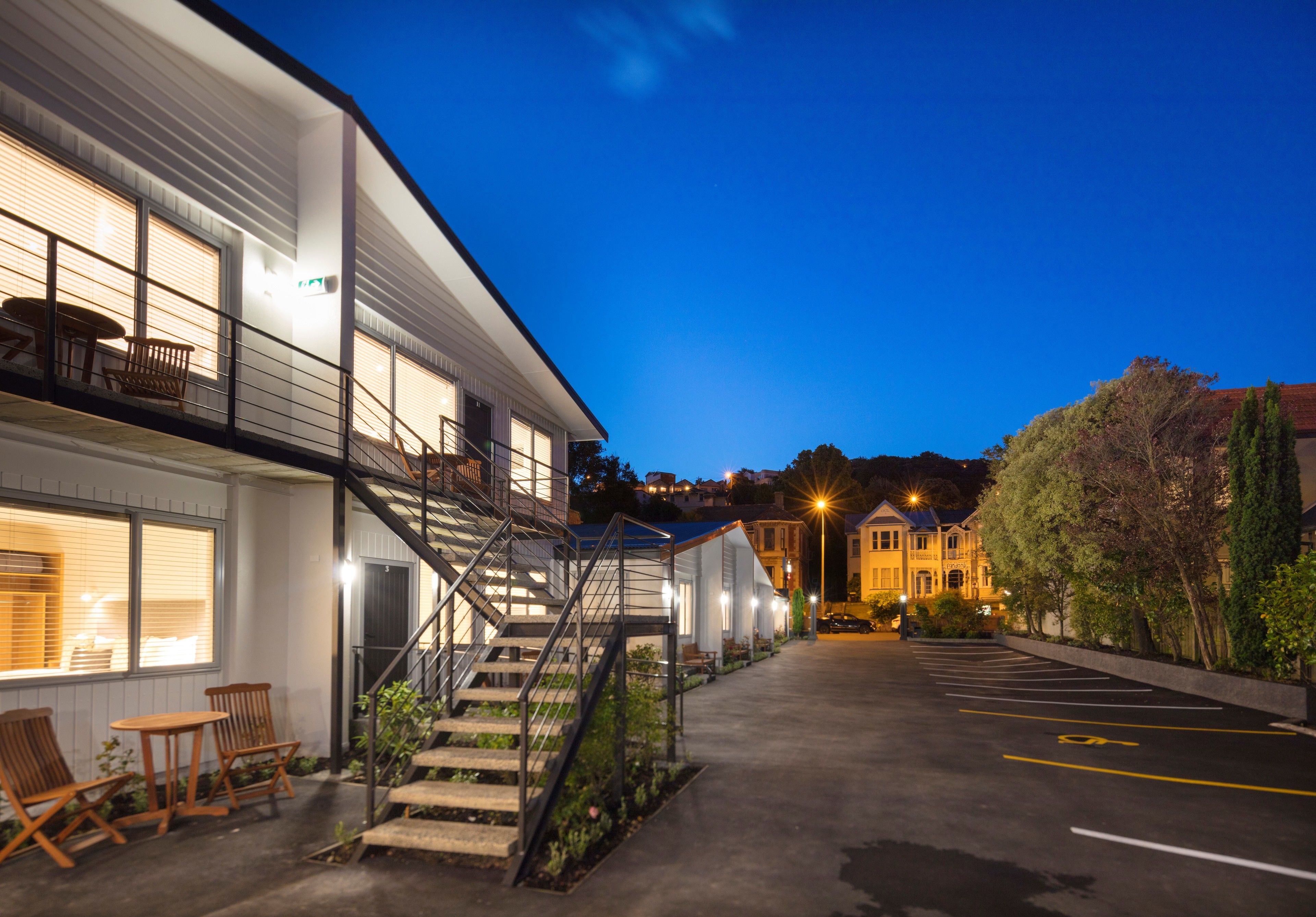 A modern building with a staircase leading to a balcony, a parking area, and a view of a hill with buildings in the background.