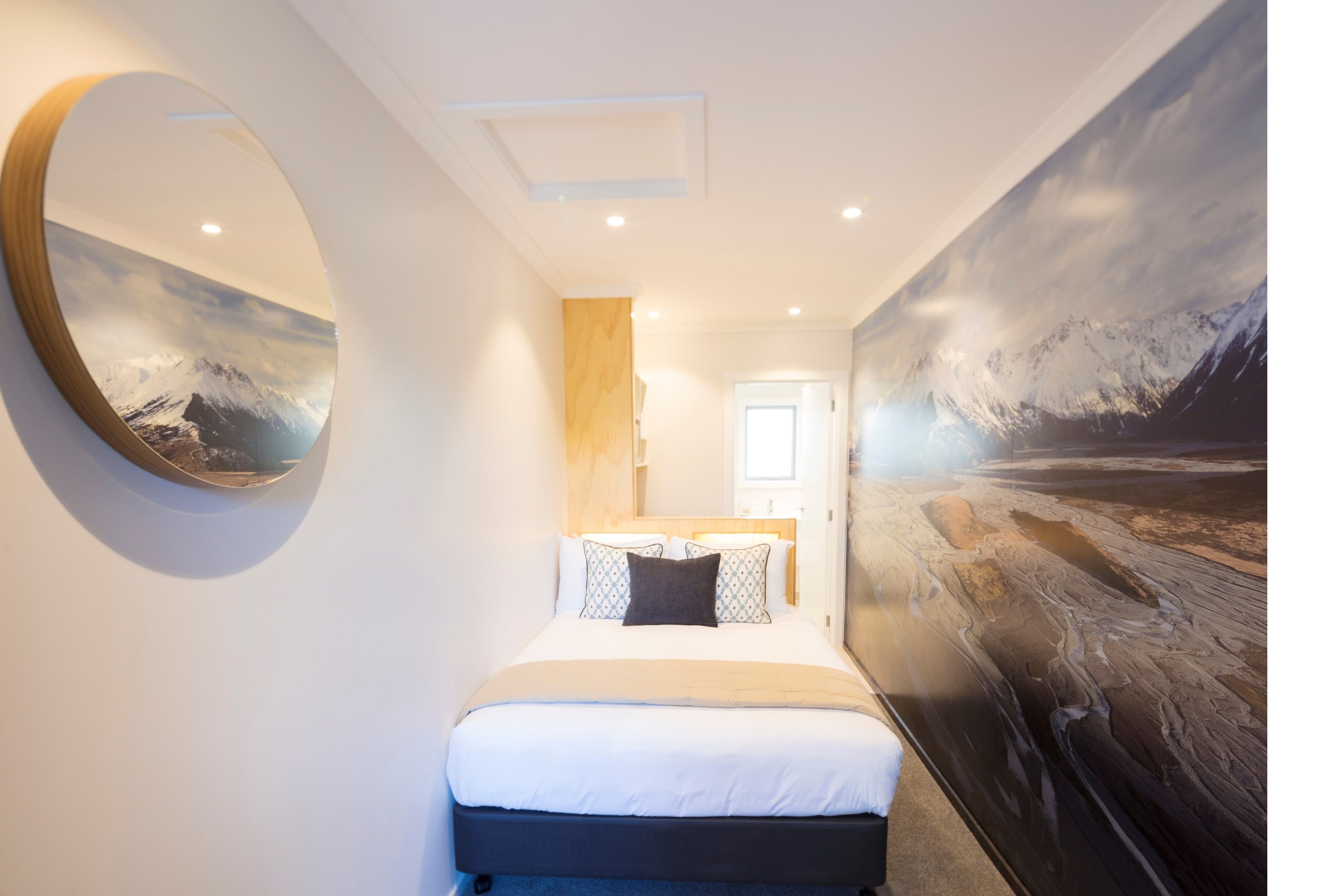 A hotel room with a large bed, a round mirror, a painting of a snowy landscape, and a wooden accent wall.