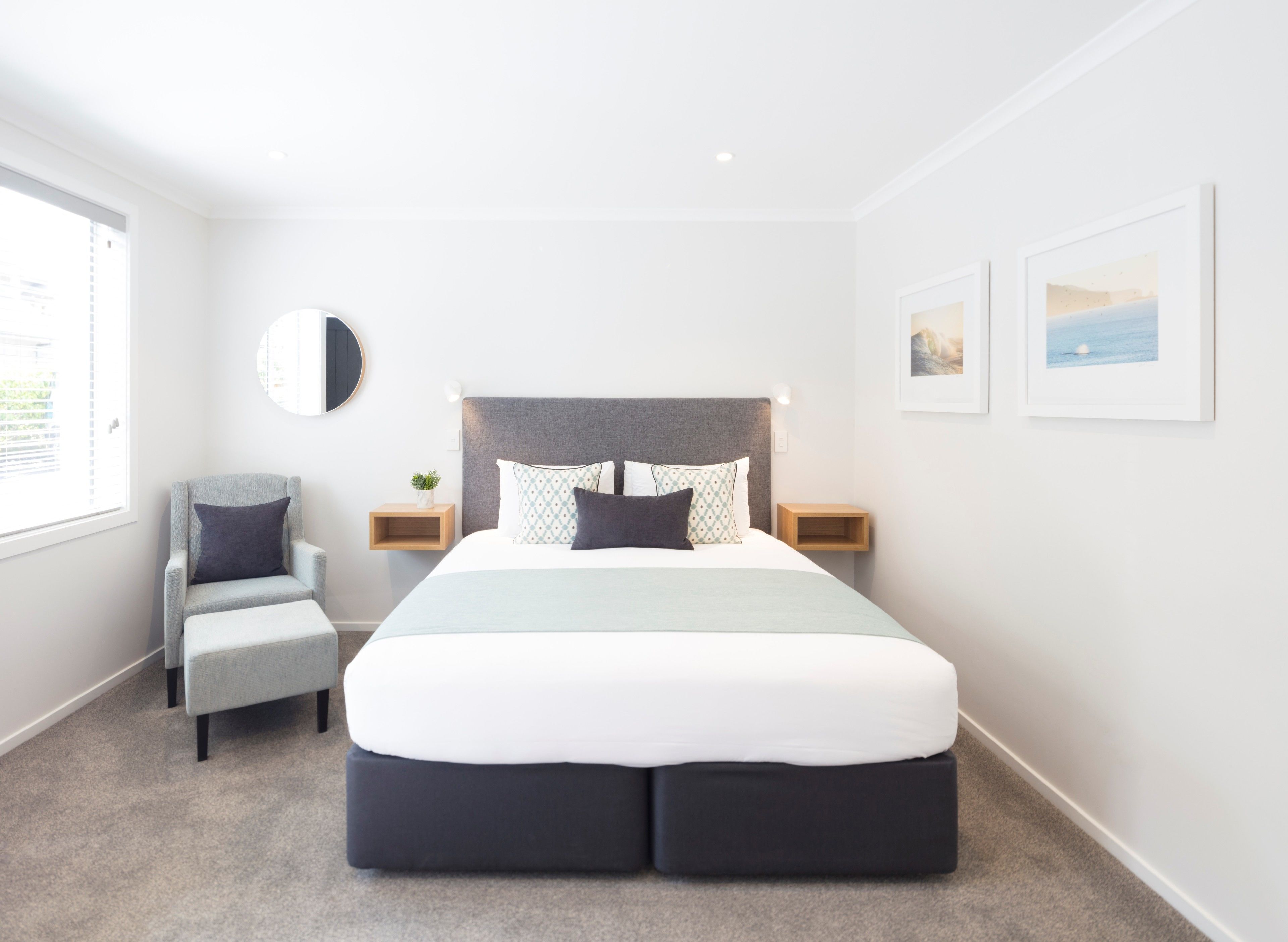A modern bedroom with a large bed, a chair, bedside tables, a round mirror, and framed pictures on the wall.
