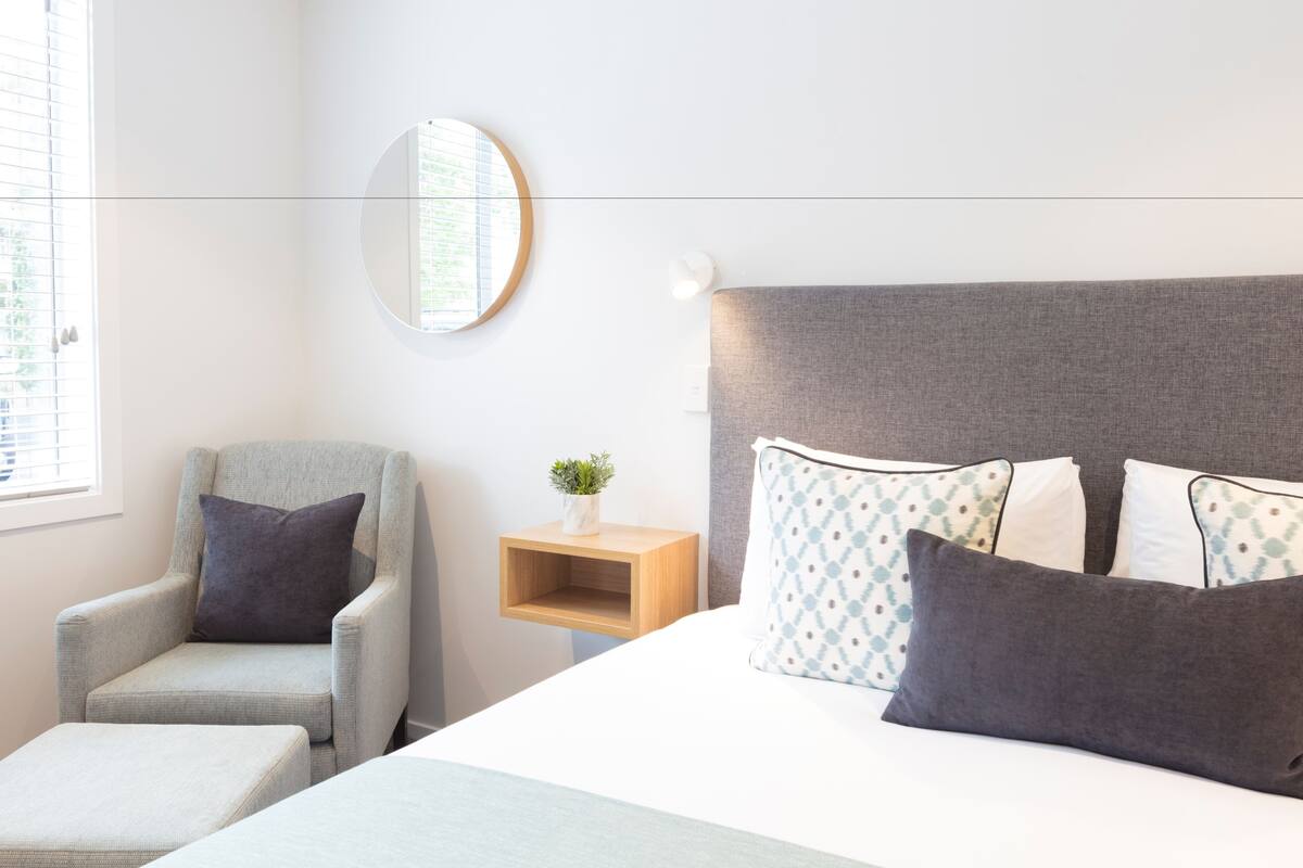 Una habitación de hotel moderna con una cama, un sillón gris, una mesita de noche de madera con una planta y un espejo redondo en la pared.