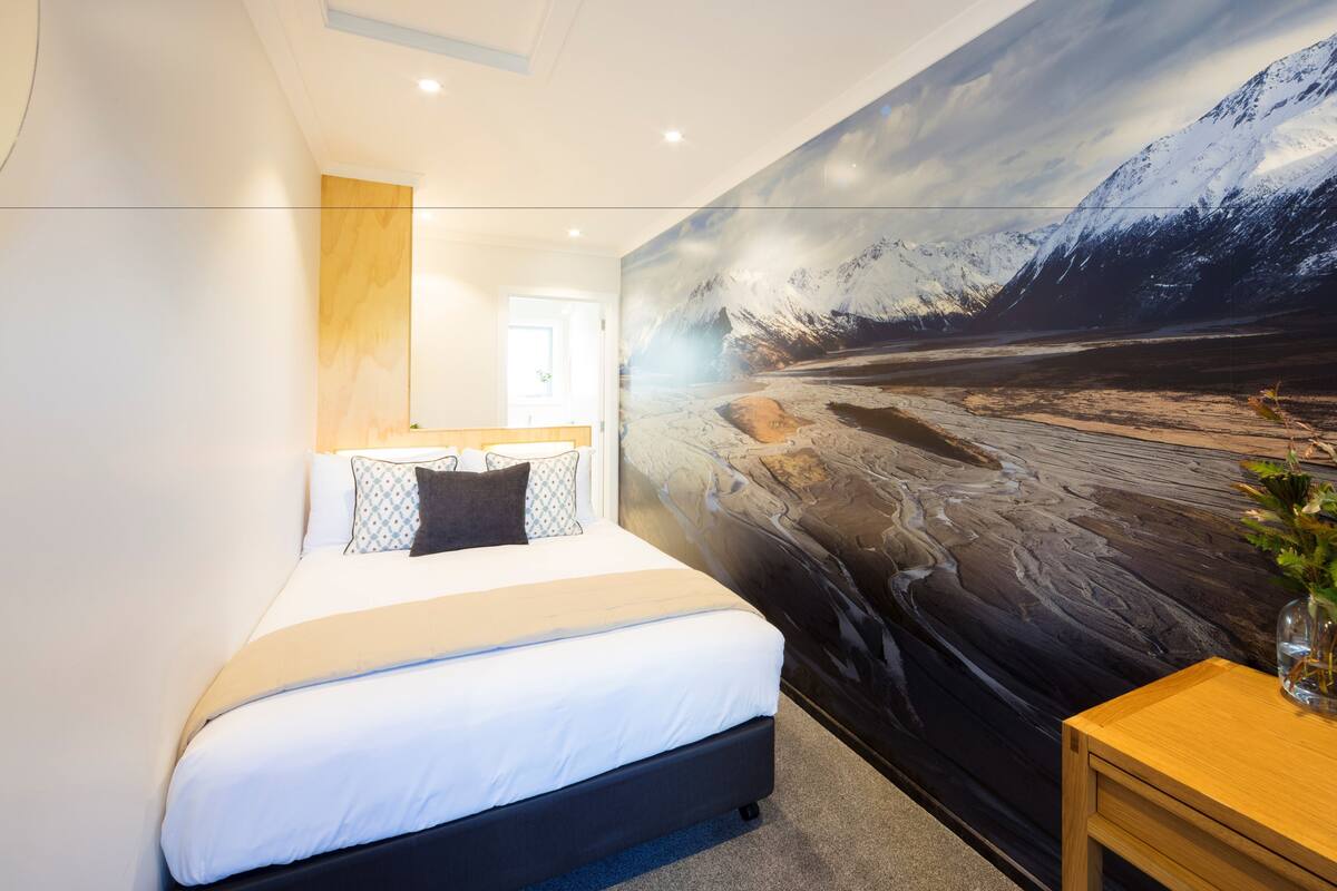 Un dormitorio moderno con una cama grande, una mesita de noche de madera y un mural en la pared con un paisaje de montaña nevada.