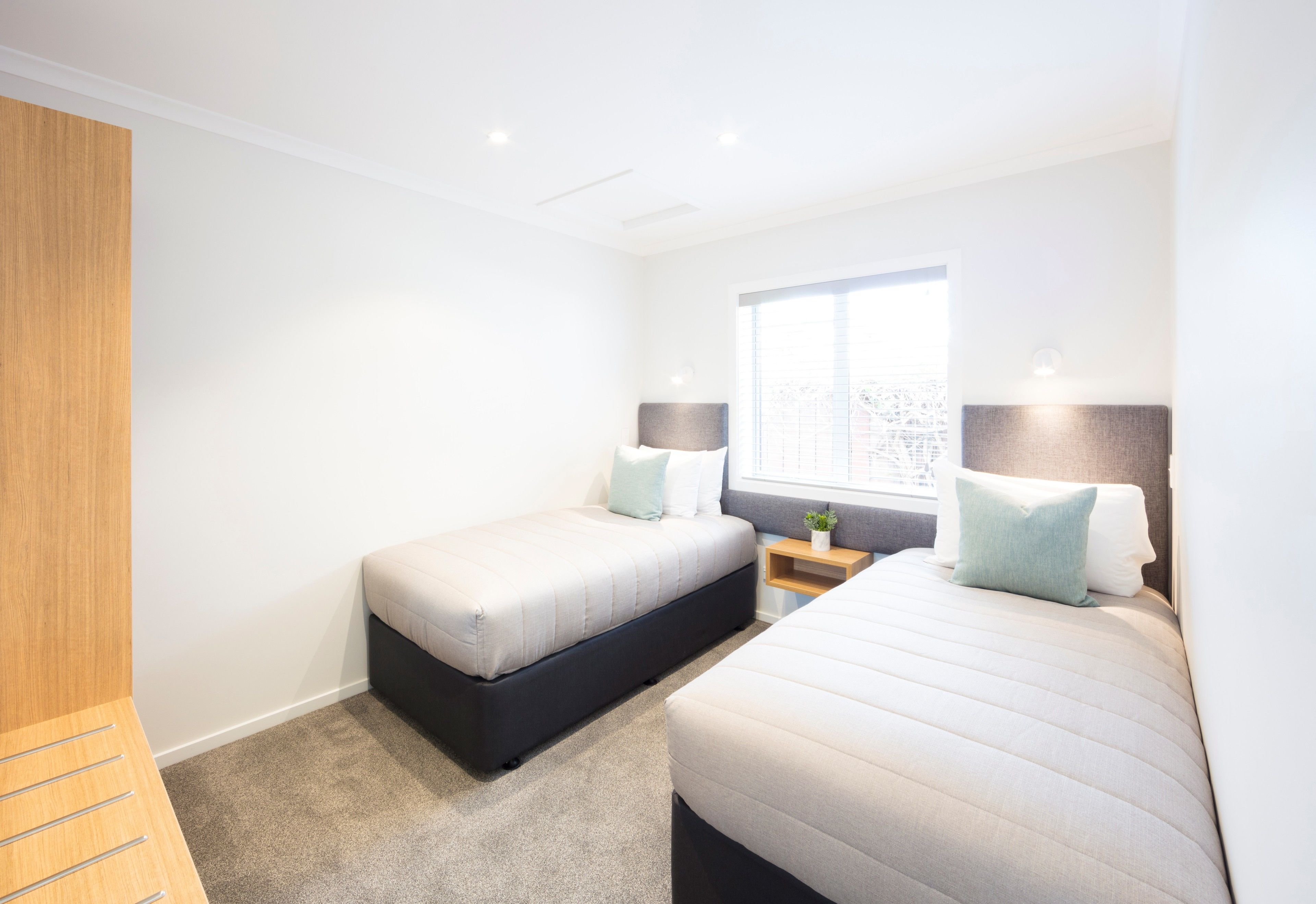 A modern hotel room with two beds, a wooden wardrobe, a small bedside table with a lamp, and a window providing a city view.