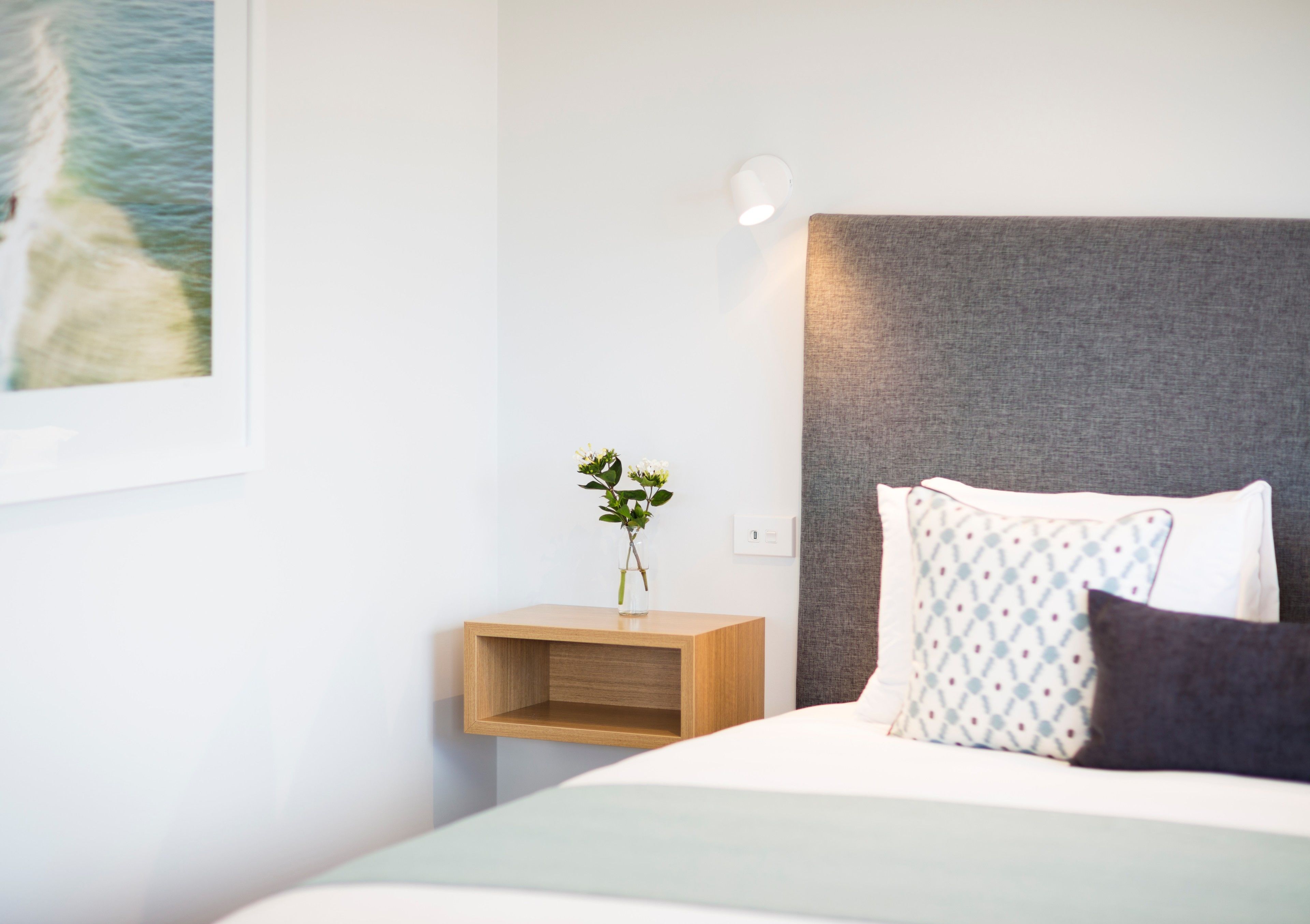 A neatly arranged hotel room with a bed, a bedside table with a vase of flowers, a wall-mounted light, and a framed picture on the wall.