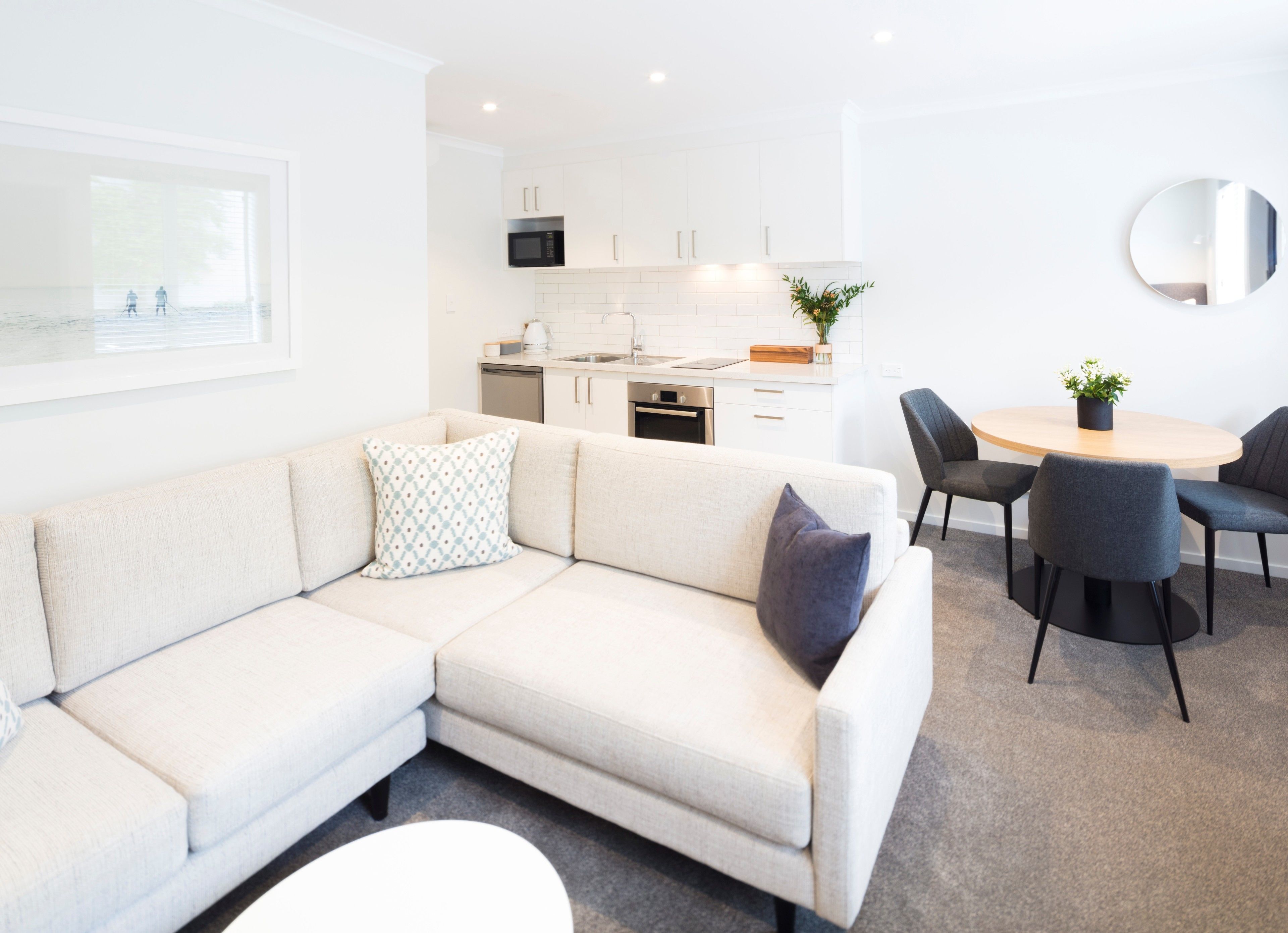 A modern living room with a beige sectional sofa, a dining table with four chairs, a kitchen area with white cabinets and a microwave, and a round mirror on the wall.