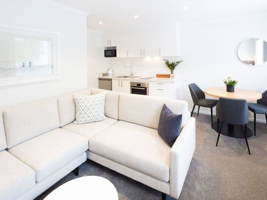 A modern living room with a beige sectional sofa, a dining table with four chairs, a kitchen area with white cabinets and a microwave, and a round mirror on the wall.