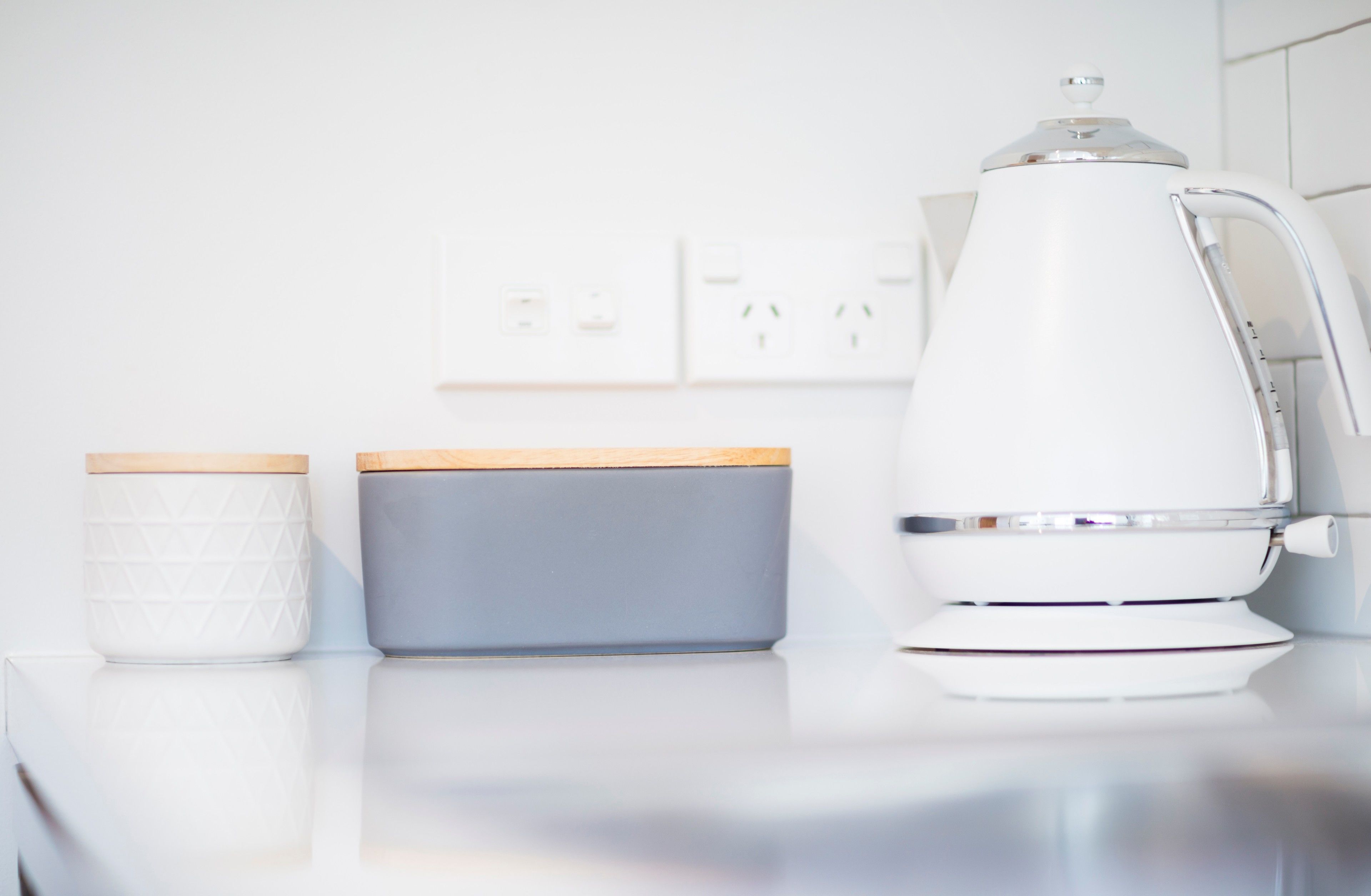 A white kitchen counter with a white kettle, a white container with a wooden lid, and a gray container with a wooden lid.