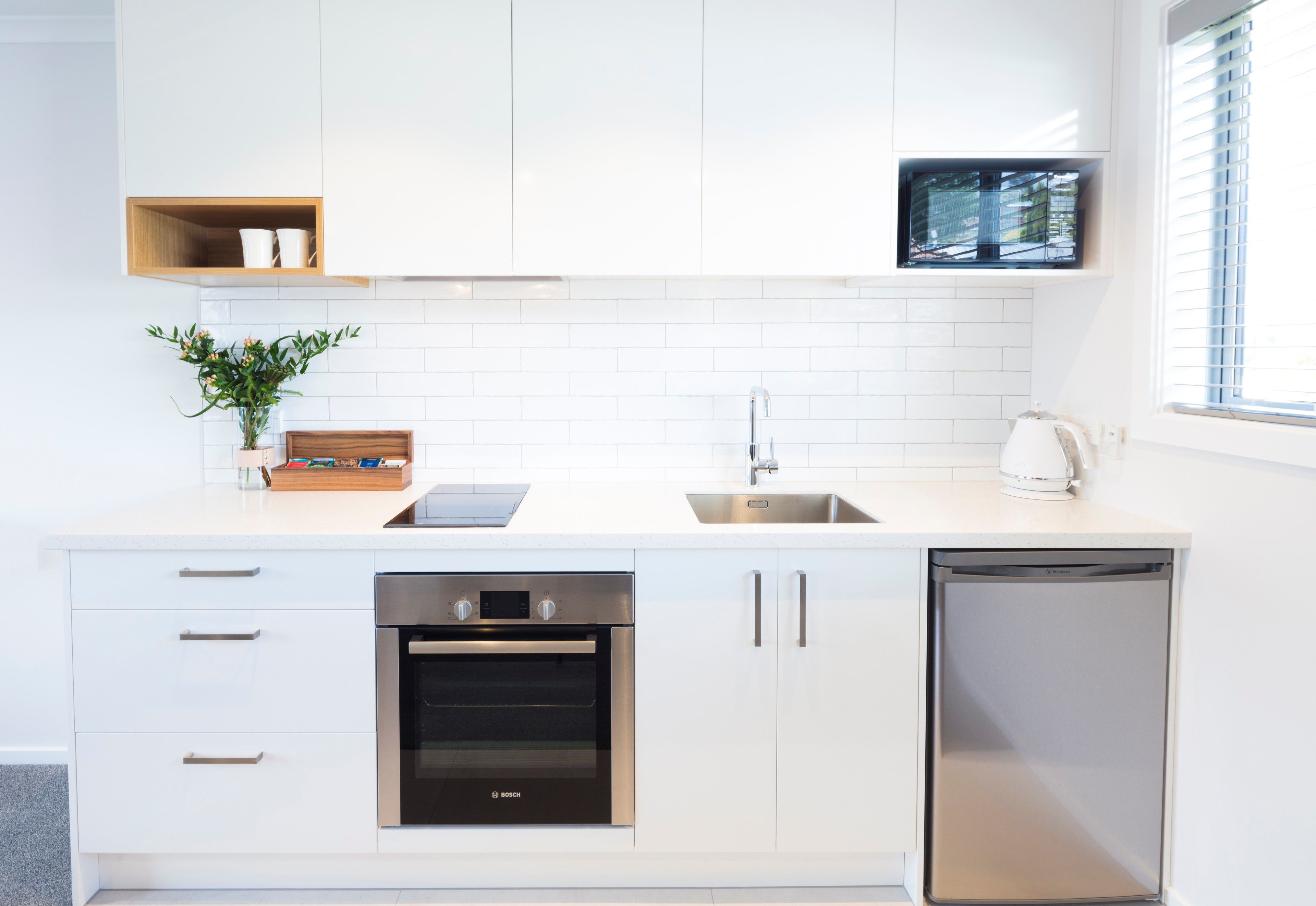 A modern kitchen with white cabinetry, a Bosch oven, a dishwasher, and a window with blinds.