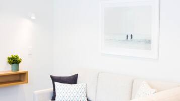 Un canapé blanc agrémenté de coussins à motifs, une petite plante posée sur une étagère en bois et un tableau encadré représentant deux personnes sur une plage.