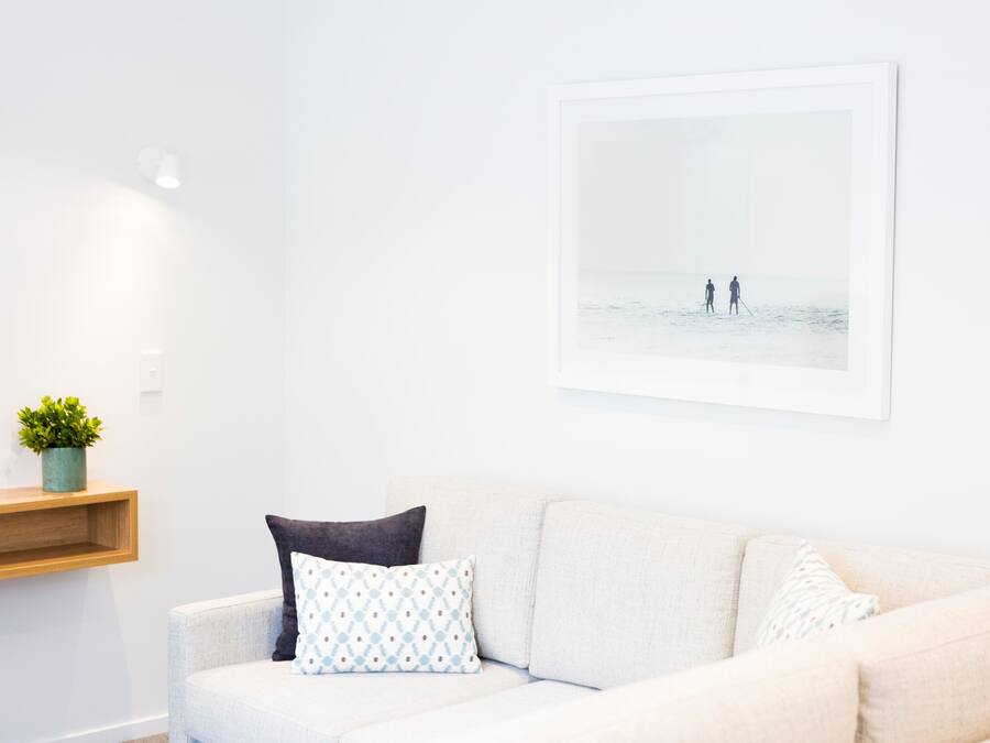 Een witte bank met patroon kussens, een klein plantje op een houten plank, en een lijst met een foto van twee mensen op het strand.