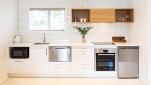 A modern kitchen with white cabinetry, a stainless steel dishwasher, an oven, and a microwave. There is a window with blinds, a shelf with kitchenware, and a potted plant. - Beechwood Boutique Accommodation (Dunedin)
