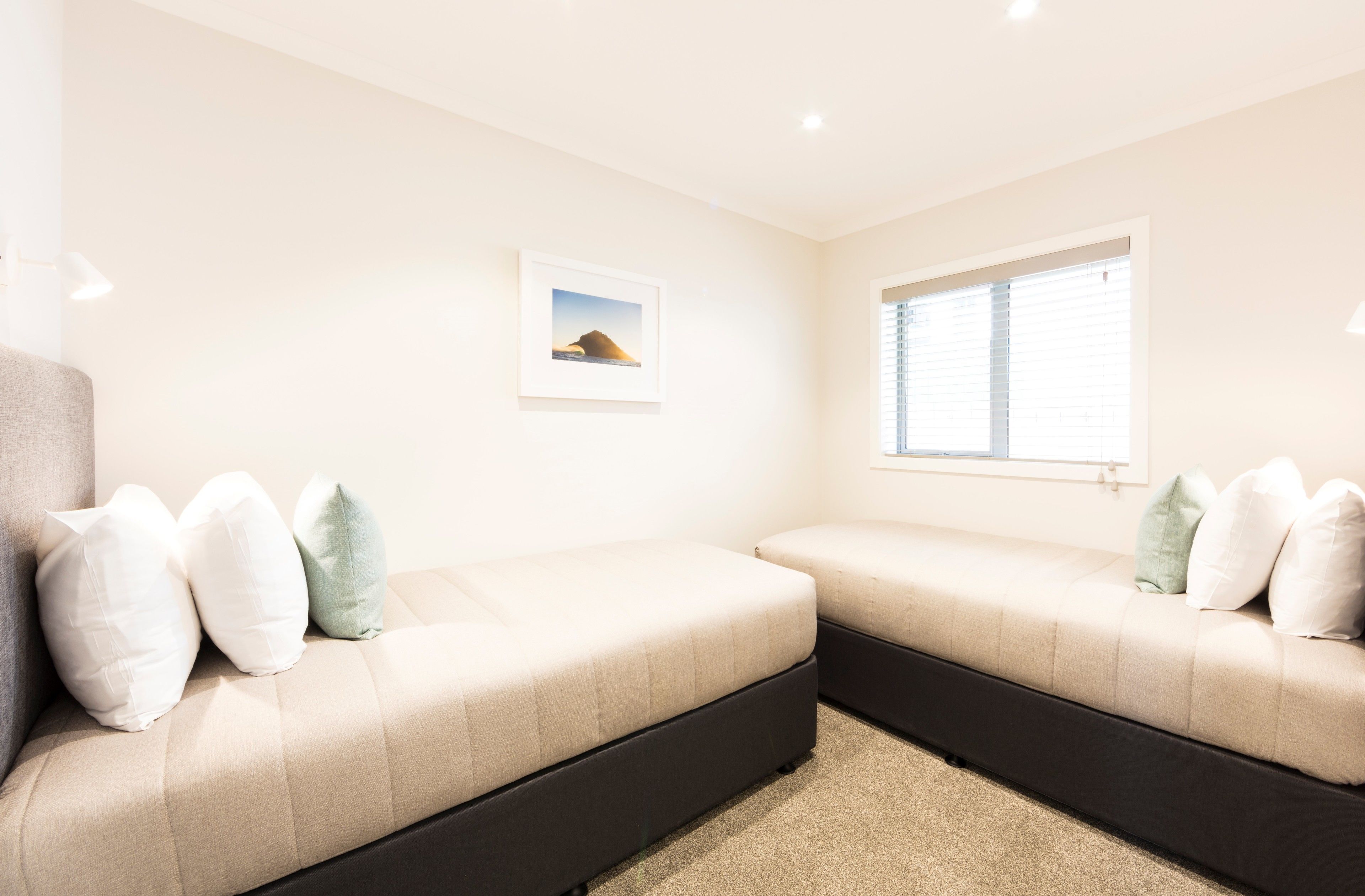 A bedroom with two beds, a window with blinds, a framed picture on the wall, and a carpeted floor.