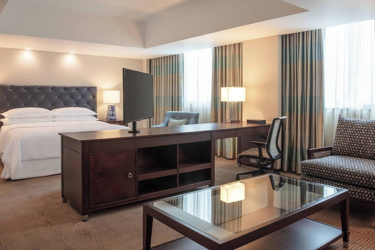 Una habitación de hotel con una cama, un escritorio con televisor, una silla, un sofá y una mesa de centro de vidrio.