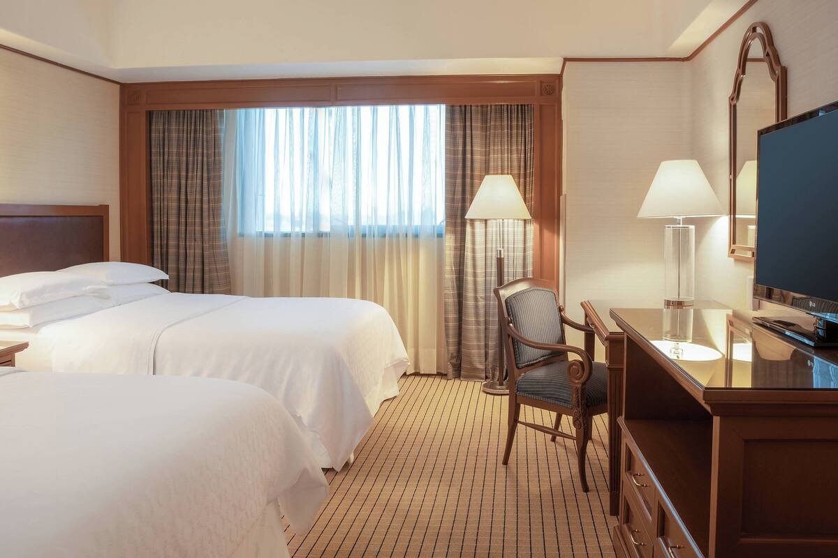 A hotel room with two beds, a desk, a chair, a TV, and a large window with curtains.