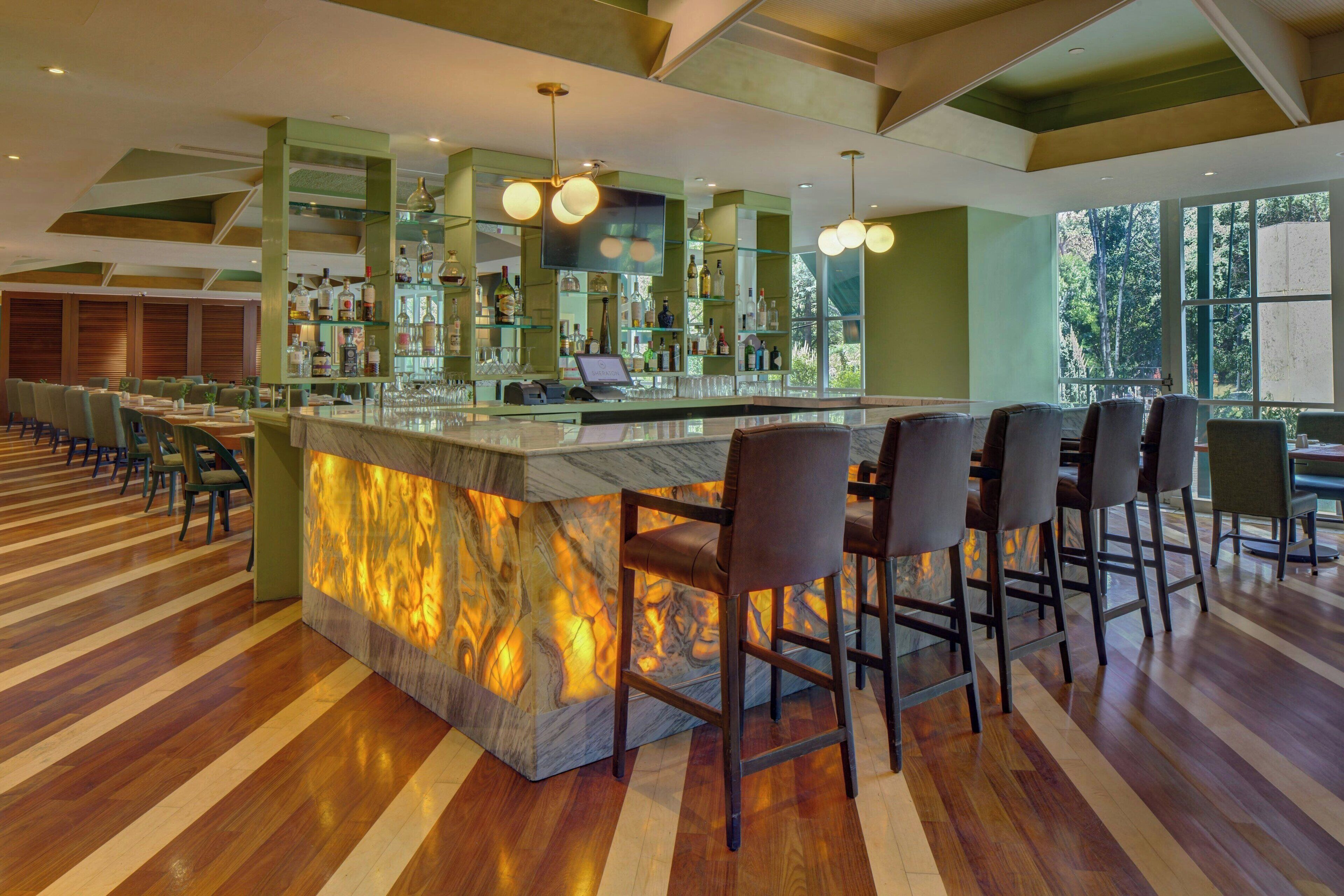 Una zona de bar con mostrador de mármol, varios taburetes y una variedad de botellas en estantes.