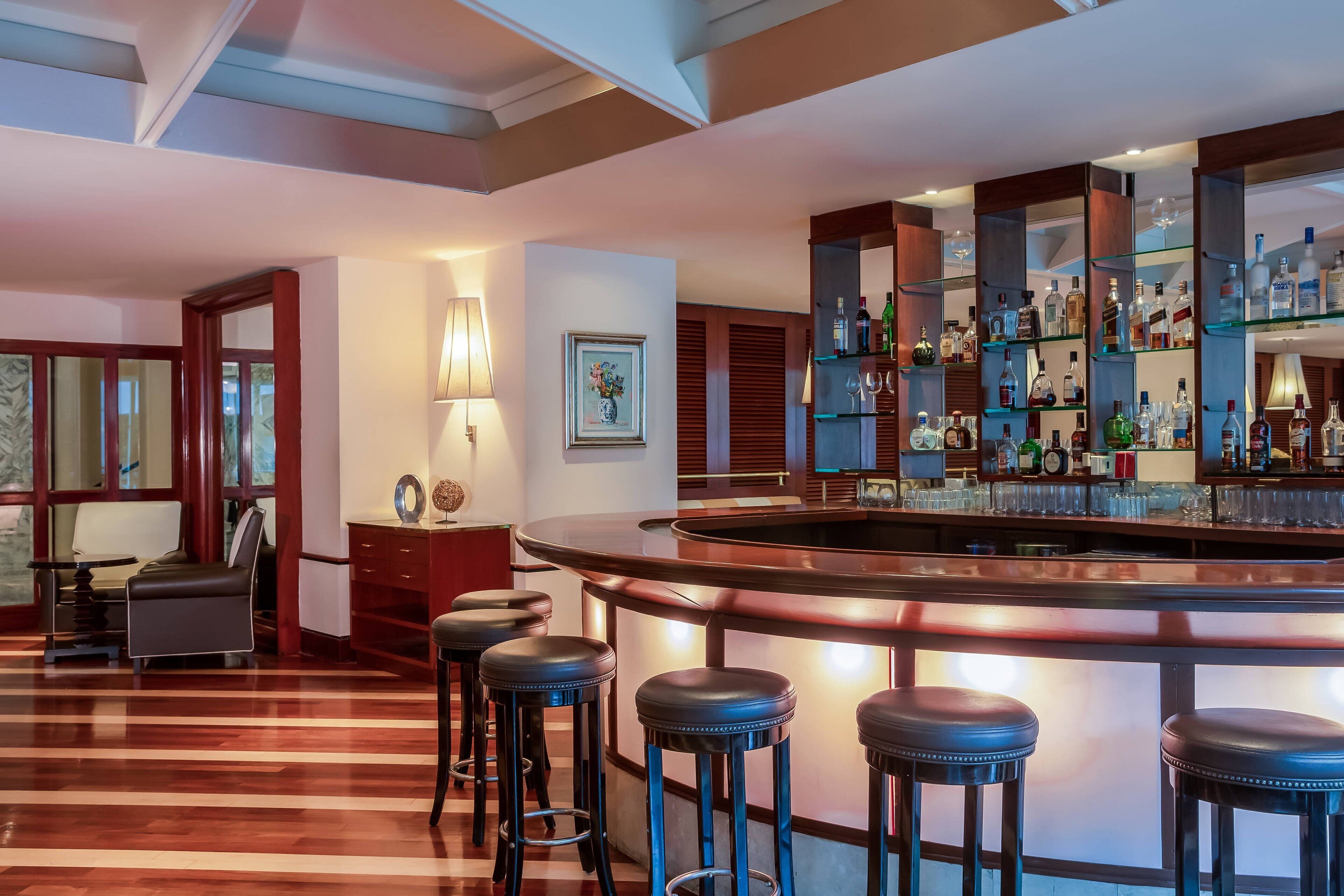 Una zona de bar con una barra curva, taburetes de madera y estantes llenos de botellas y vasos.