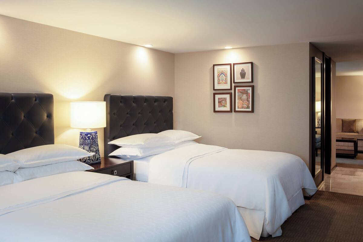 Habitación de hotel con dos camas, una mesita de noche, una lámpara y cuadros en la pared.