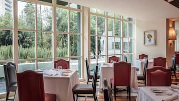 Ein Restaurant mit großen Fenstern, Tischen mit weißen Tischdecken und roten Stühlen.