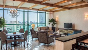 A modern hotel room with a large window, a round dining table, a sofa, a TV, and a bar area.