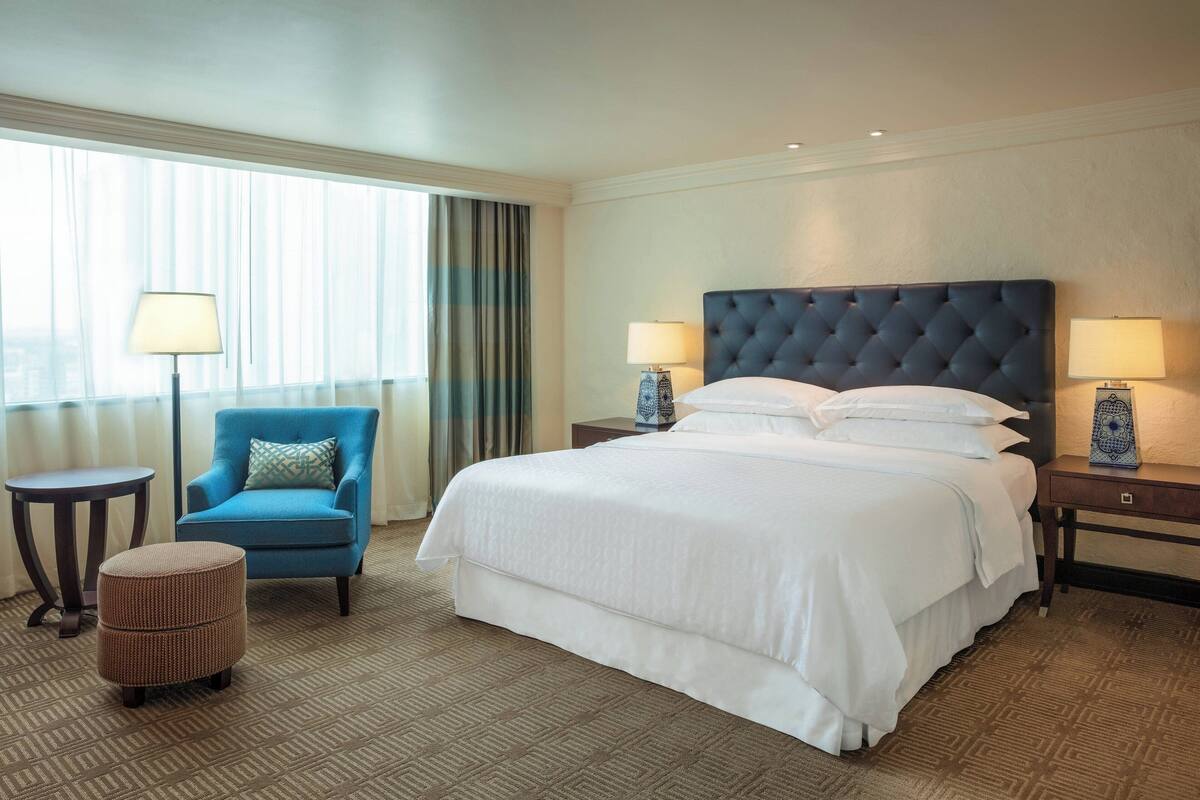 Habitación de hotel con una cama grande, un sillón azul, un pequeño reposapiés, dos mesitas de noche con lámparas y vistas a través de cortinas translúcidas.