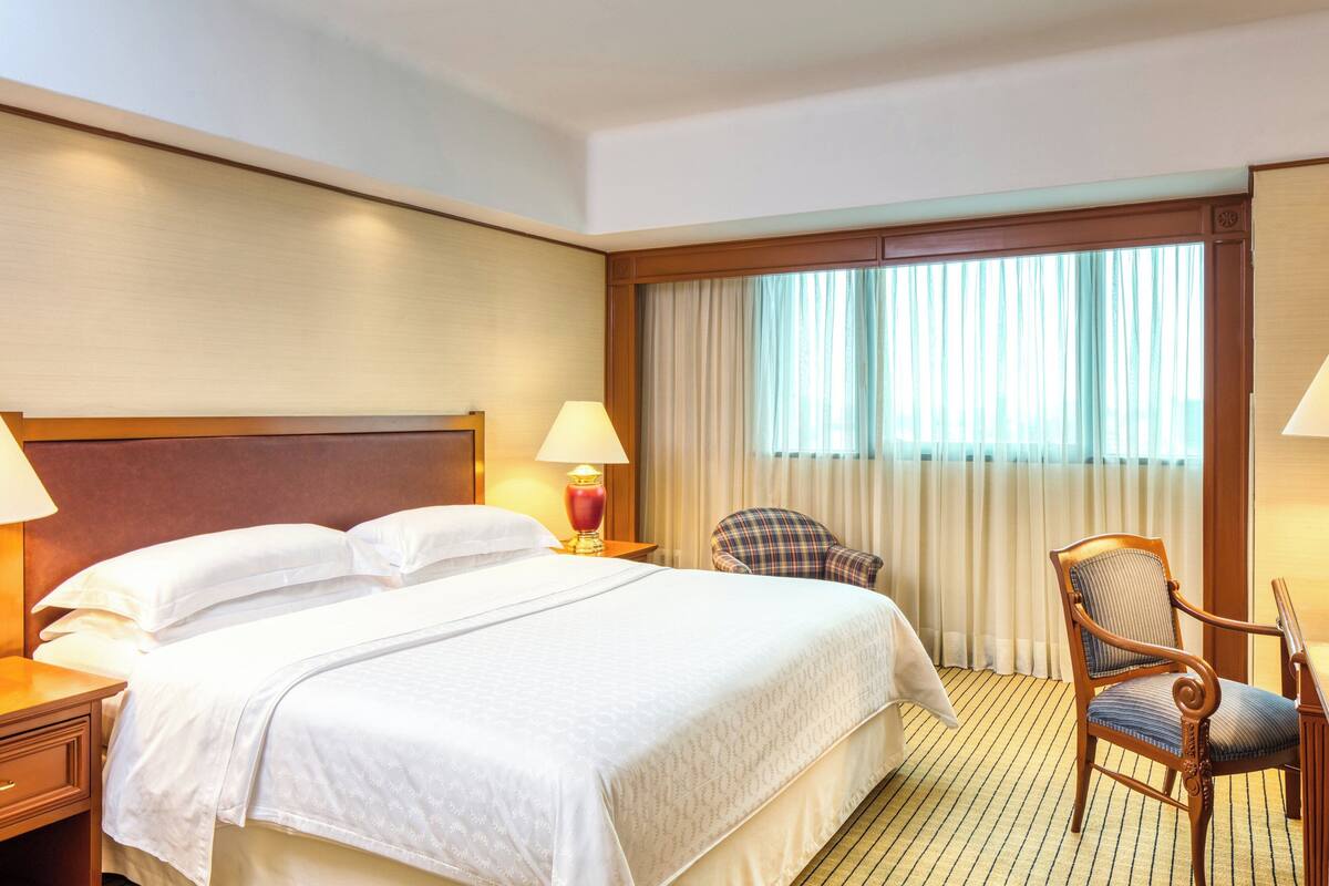 A hotel room with a large bed, a chair, a nightstand, a lamp, and a window with curtains.