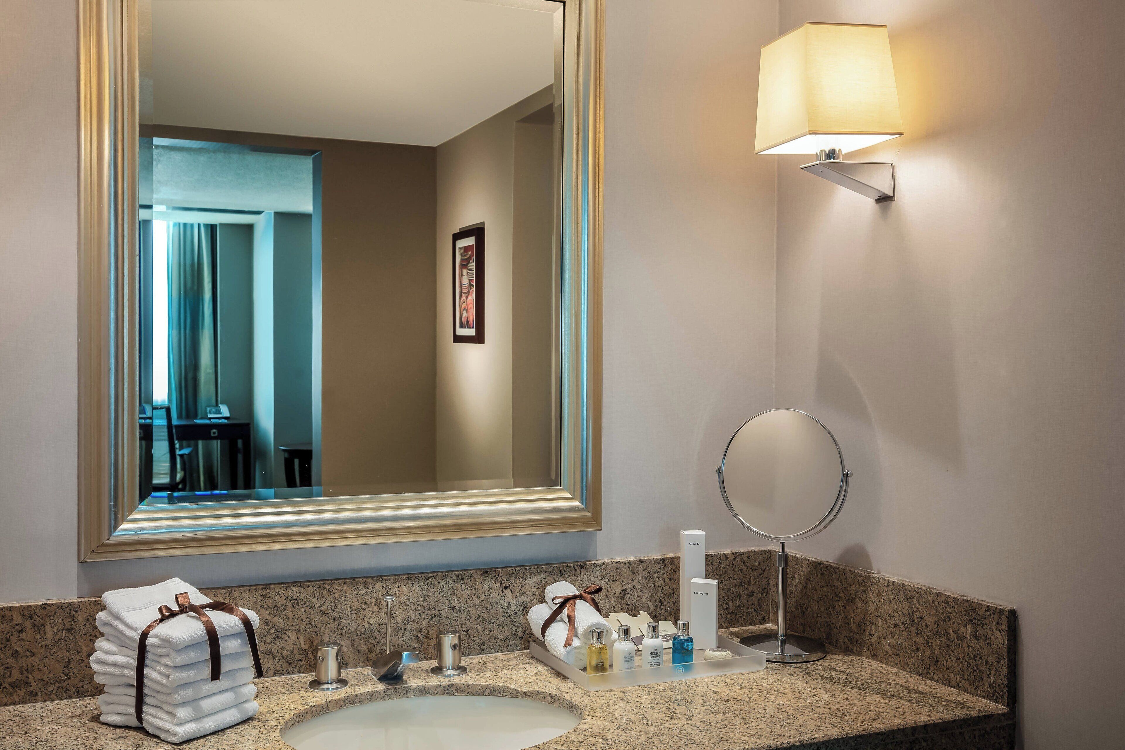 Un baño de hotel con un espejo grande, un lavamanos, un perchero para toallas, un espejo redondo y una lámpara de pared.