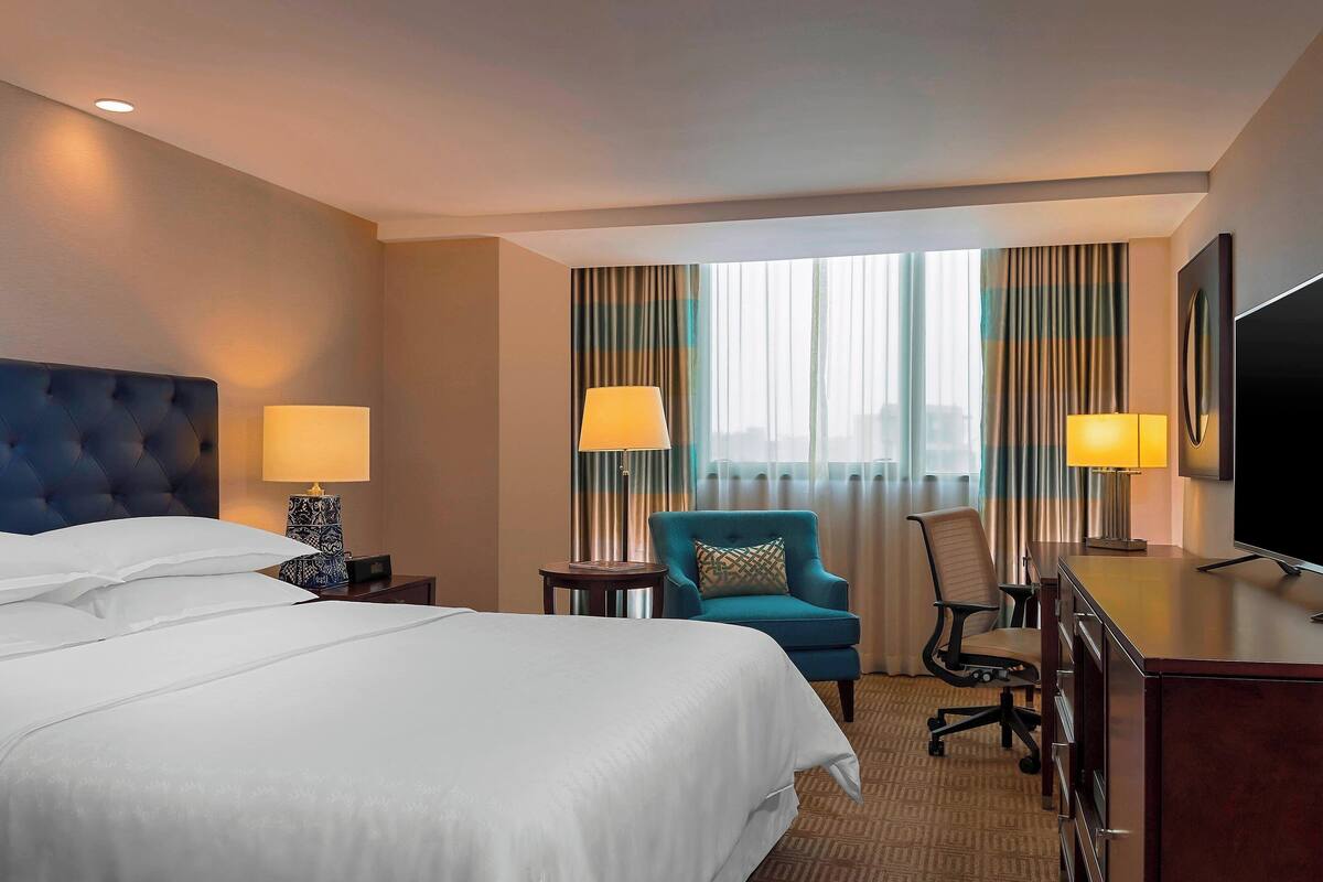 A hotel room with a large bed, two bedside tables with lamps, a desk with a television, a blue armchair, and a view of the city through large windows.