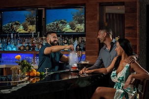 Una zona de bar con un cantinero preparando una bebida, un hombre y una mujer sentados en la barra, y un televisor mostrando una escena submarina.