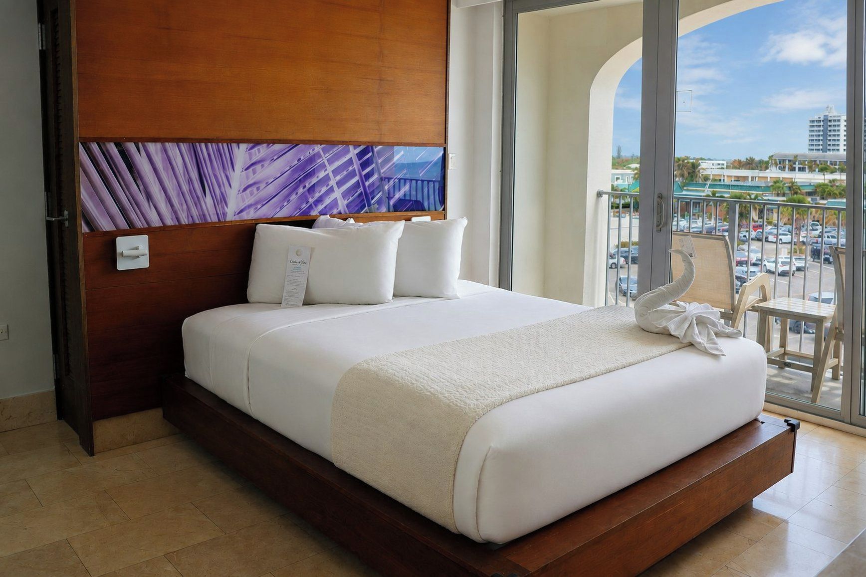 Habitación de hotel con una cama grande, cabecera de madera, vista de la ciudad desde un balcón y un cuadro sobre la cama.