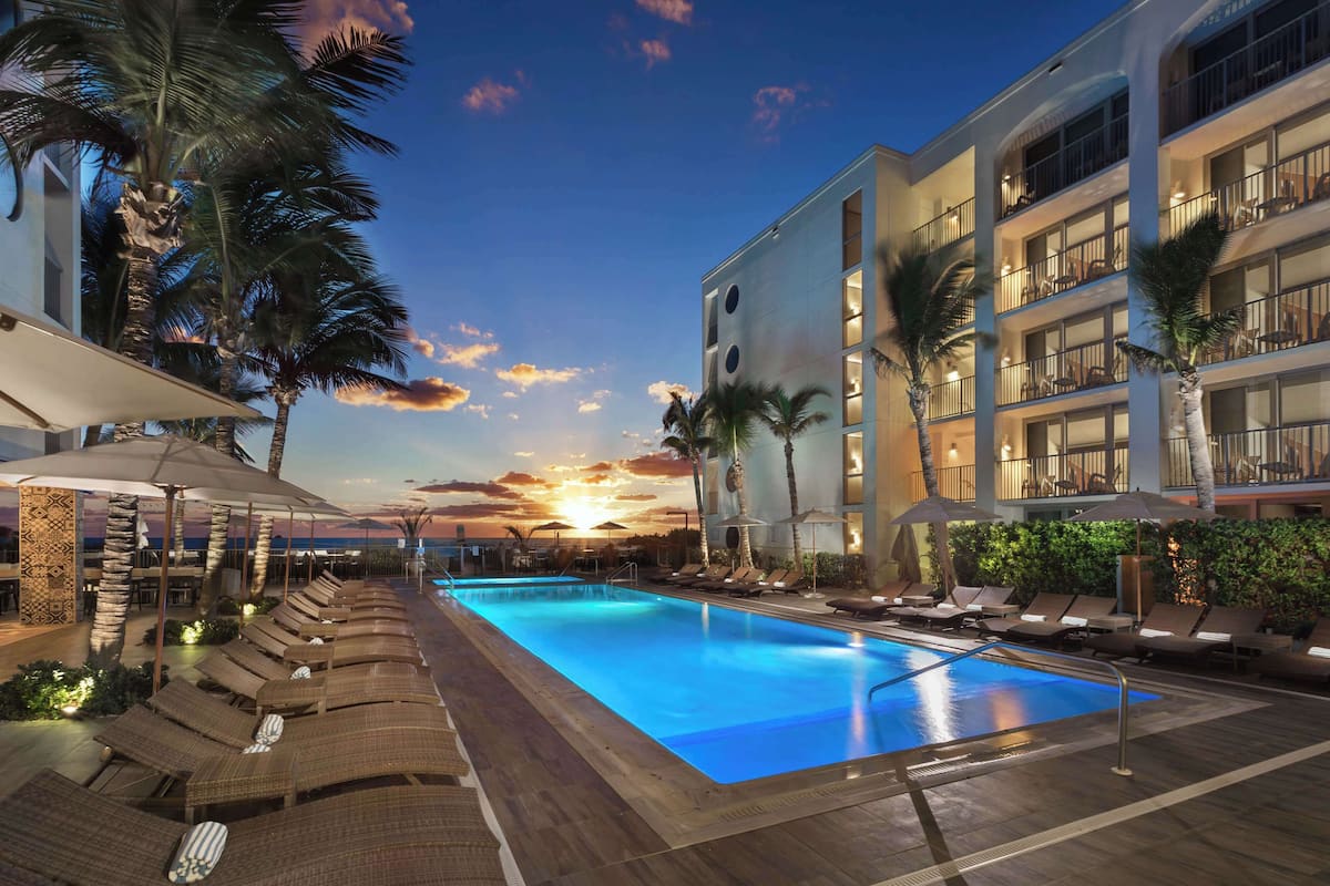 A swimming pool with lounge chairs and umbrellas, surrounded by palm trees and a building with balconies.