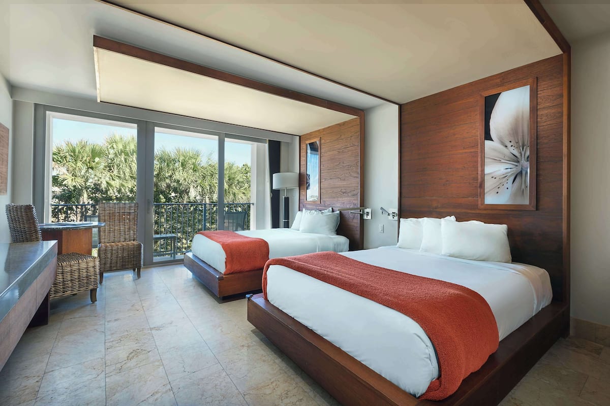 A modern hotel room with two beds, a balcony, wicker furniture, and a large window.