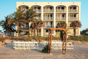 Un hotel frente a la playa con varios balcones, una playa de arena y sillas blancas dispuestas para un evento.