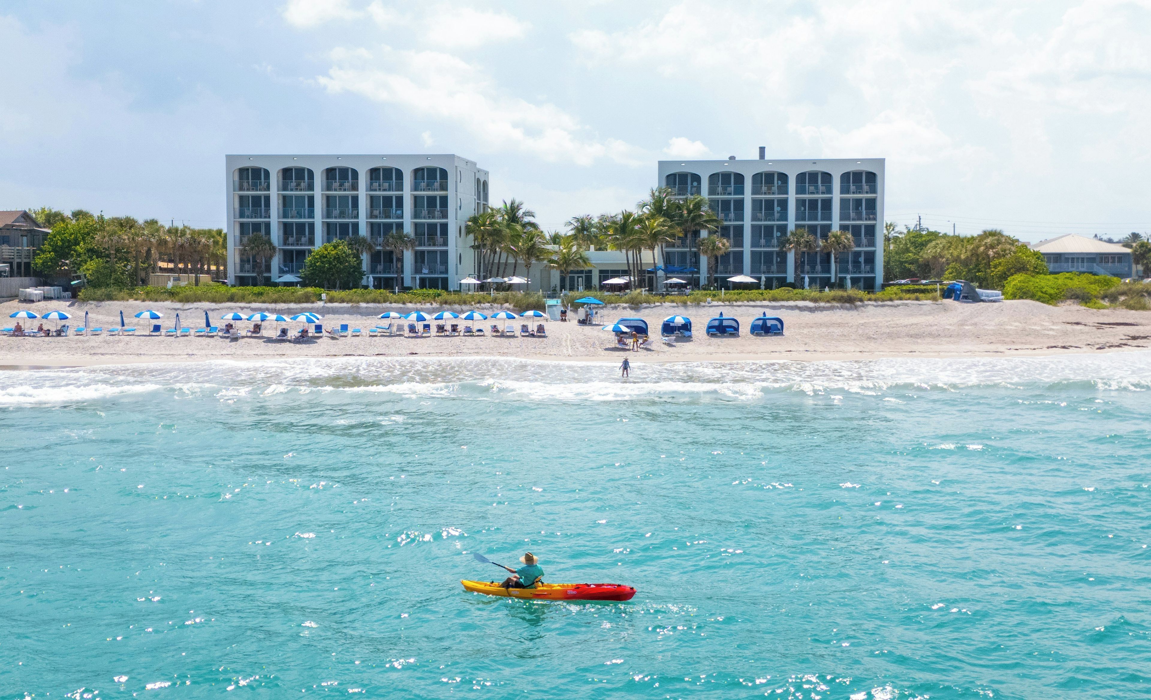 A beach with a kayaker, beach umbrellas, and two large buildings in the background.