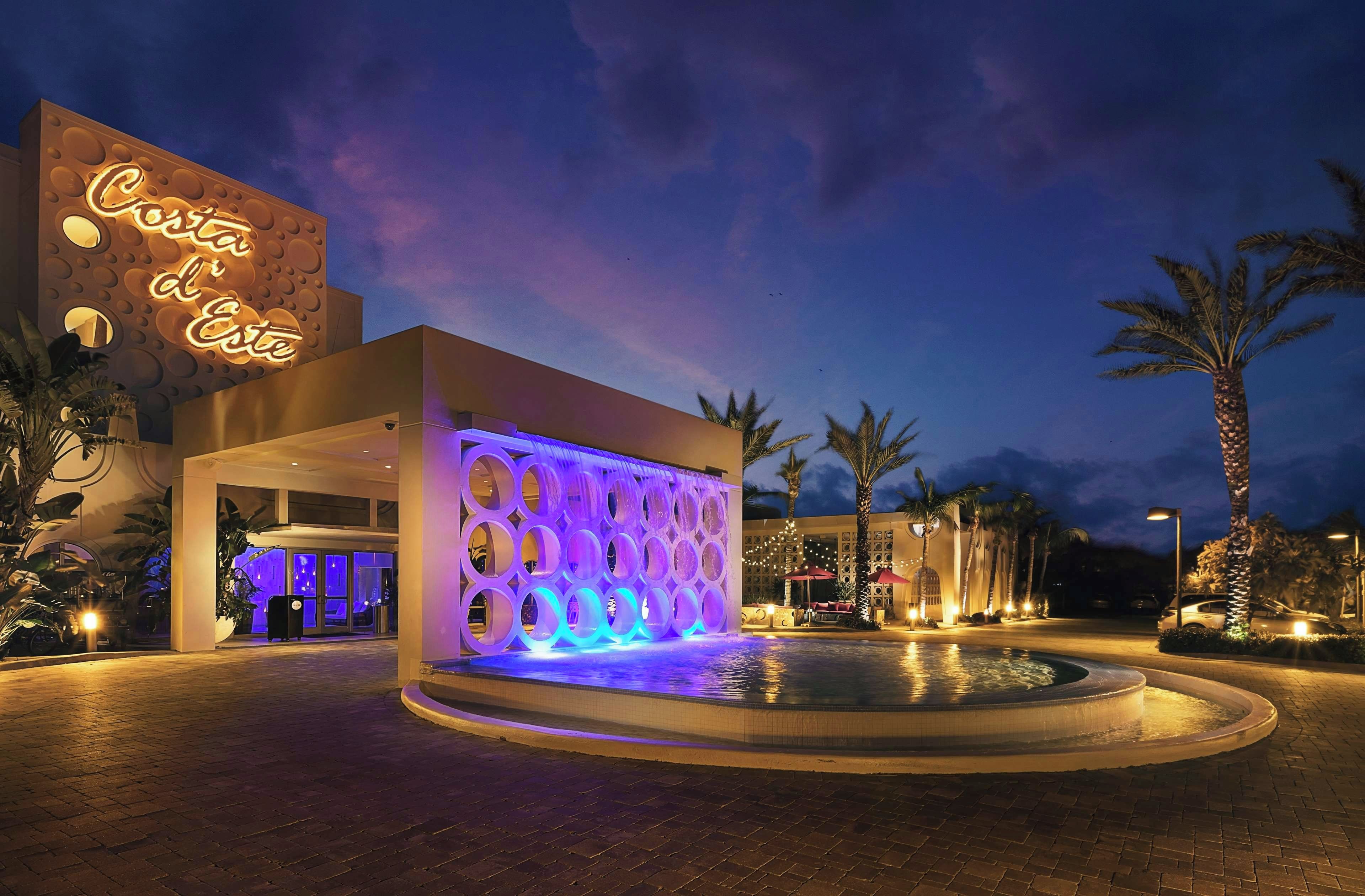 A hotel entrance with a pool, palm trees, and a neon sign.
