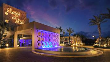 A hotel entrance with a pool, palm trees, and a neon sign.