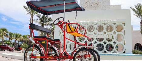 Un vehículo rojo de tres ruedas con toldo, estacionado frente a un edificio con un muro decorativo.