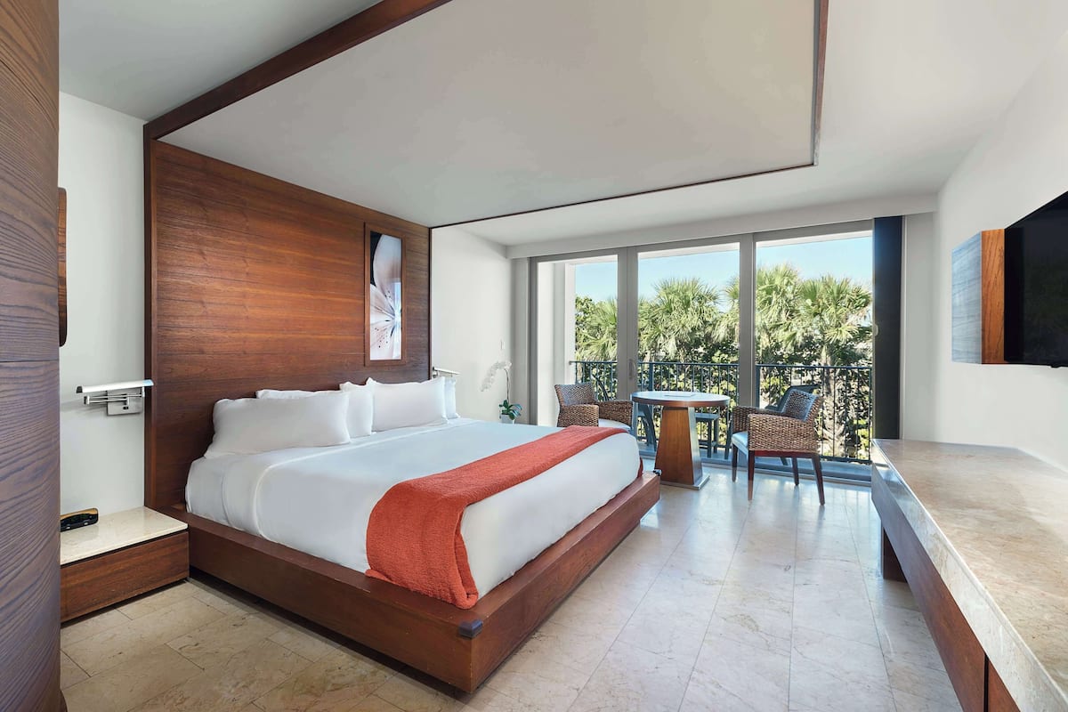 A modern hotel room with a large bed, a balcony with outdoor seating, and a large window offering a view of palm trees.