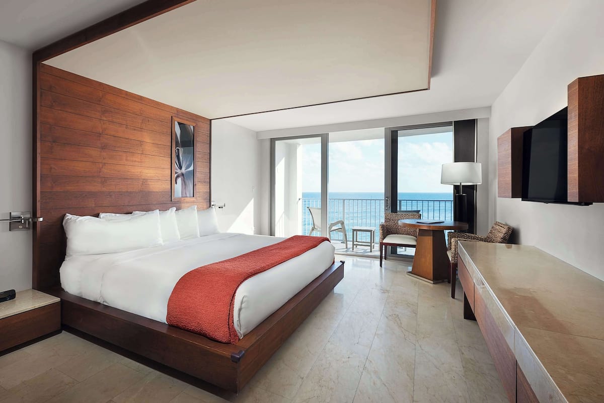 A modern hotel room with a large bed, a desk, a chair, and a view of the sea.